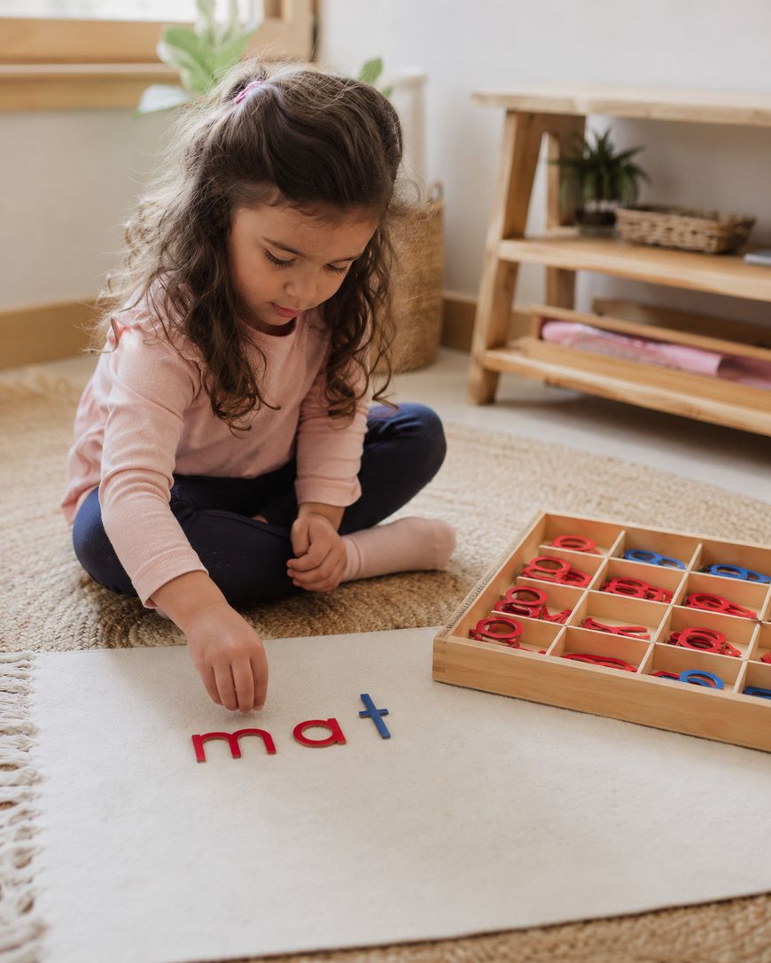 A young girl sits on the floor arranging colored wooden letters to spell mat on a white rug, with a box of more letters beside her in a cozy, sunlit room.