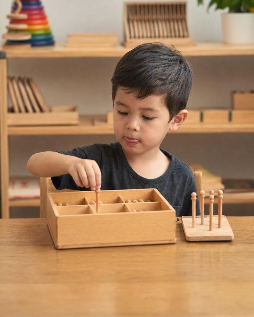 A young boy sits at a table, focused on a wooden Montessori math toy with rods and counters, sorting or placing pieces into a box with compartments. Shelves with educational materials are in the background.