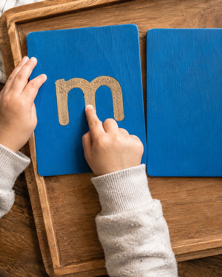 A child traces a textured lowercase letter m on a blue wooden card with their finger, using their other hand to hold the card steady on a wooden tray.