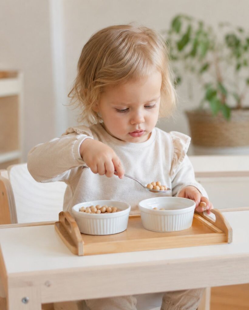 A young child with blonde hair sits at a table, holding a spoon and eating chickpeas from a bowl. There are two white bowls on a wooden tray in front of the child. A green plant is visible in the background.