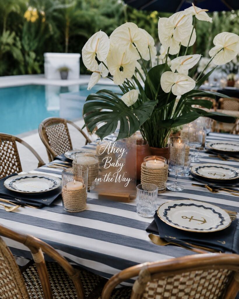 Outdoor table set for a nautical-themed baby shower, with striped tablecloth, anchor plates, candles, tropical white flowers, and a sign reading Ahoy, Baby on the Way beside a swimming pool.