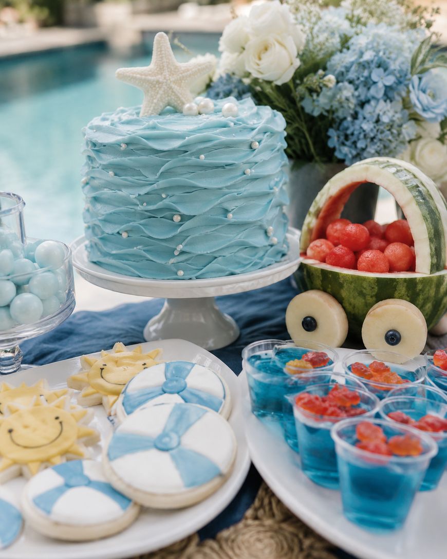 A summer-themed dessert table by a pool with a blue cake topped with a starfish, sun and beach ball cookies, blue jelly cups with gummy bears, a watermelon bowl with fruit, and a bouquet of white and blue flowers.