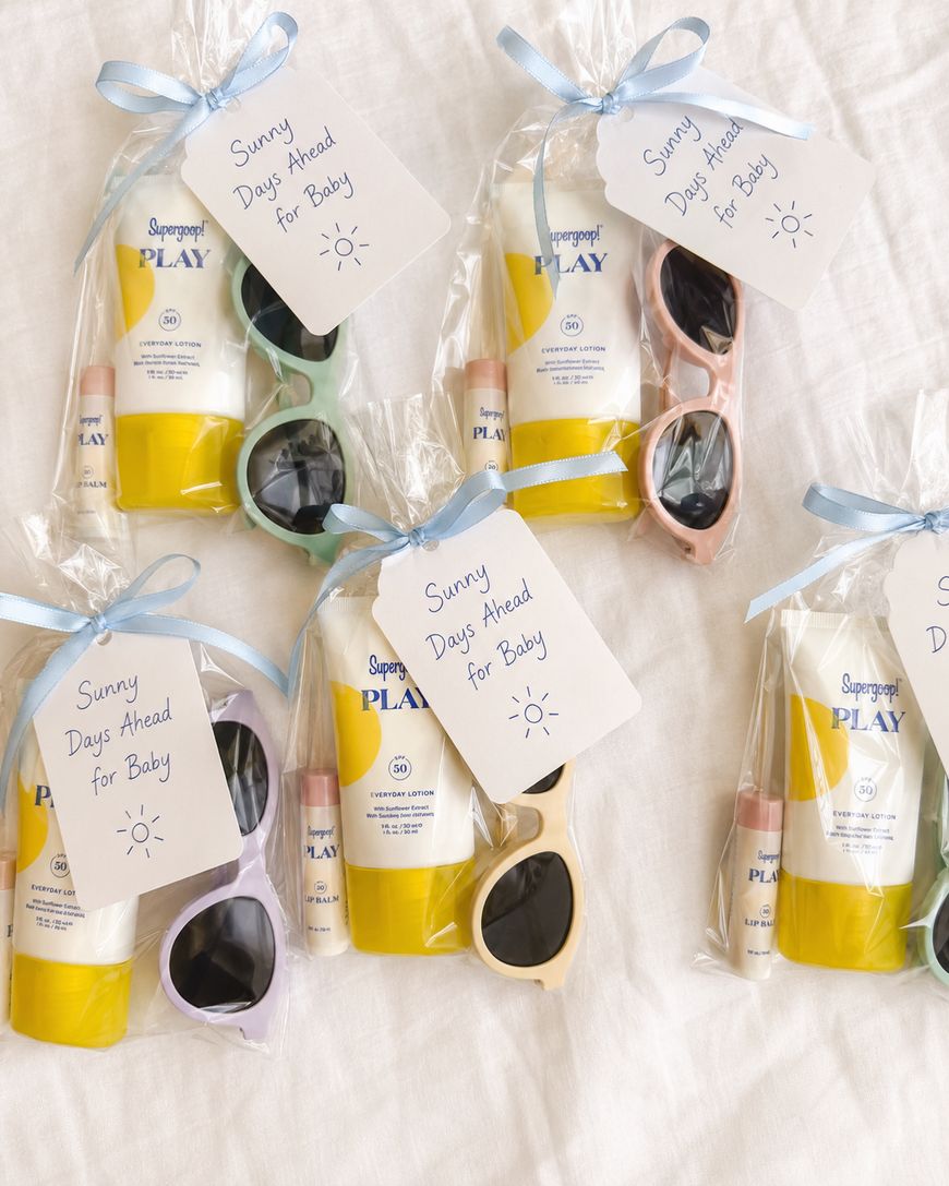 Five gift bags with sunglasses, sunscreen, and lip balm are displayed on a white cloth. Each bag is tied with a ribbon and a tag that reads Sunny Days Ahead for Baby.