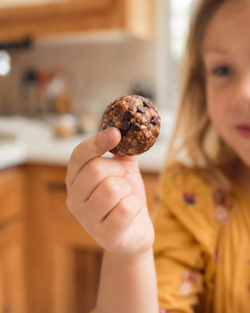 A young child in a yellow shirt holds up a round chocolate chip energy bite in a kitchen, with the treat in focus and the childs face blurred in the background.