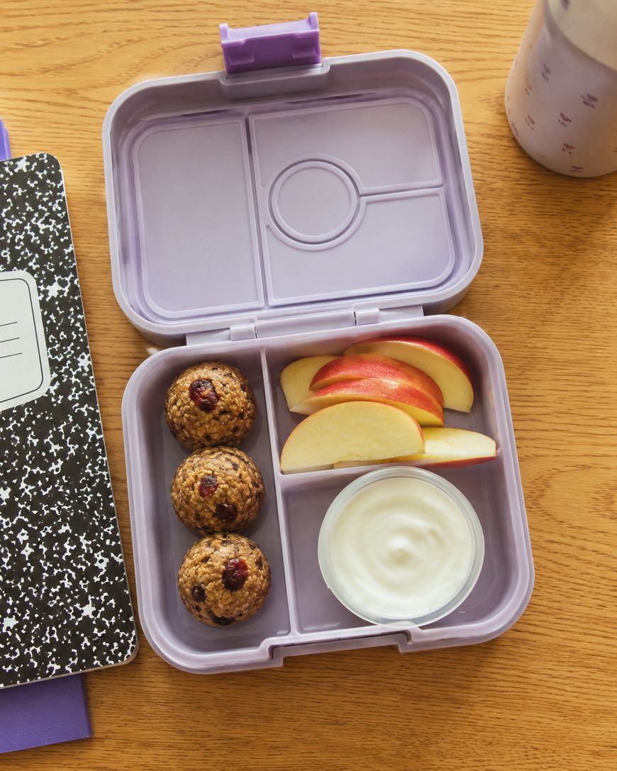 A purple lunchbox with compartments holding three baked oatmeal bites, sliced red apple, and a cup of yogurt sits on a wooden table next to a black and white notebook and a partially visible tumbler.