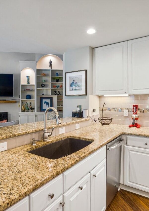 A kitchen with white cabinets, granite countertops, and a stainless steel sink and dishwasher. A bar area with shelves and decorative items is in the background.