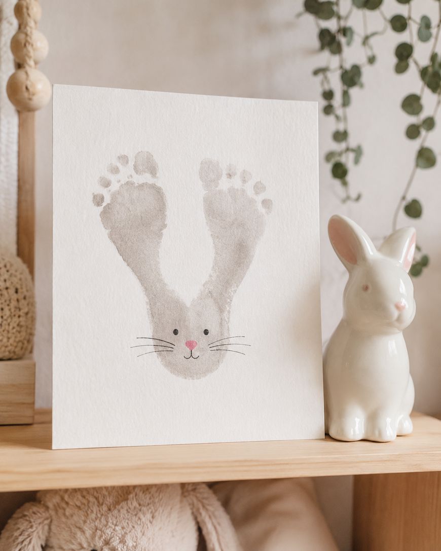 A card with a bunny face drawn using two gray footprints for ears, with added whiskers, eyes, and a pink nose, is displayed on a wooden shelf next to a white ceramic rabbit and some green hanging plants.