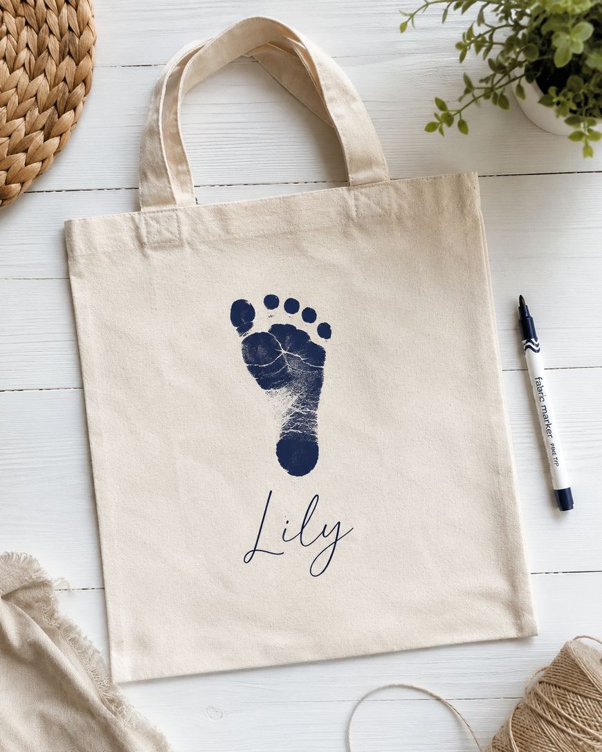 A beige tote bag with a navy blue baby footprint and the name Lily written in cursive below it, placed on a white wooden surface with a marker, plant, and woven mat nearby.
