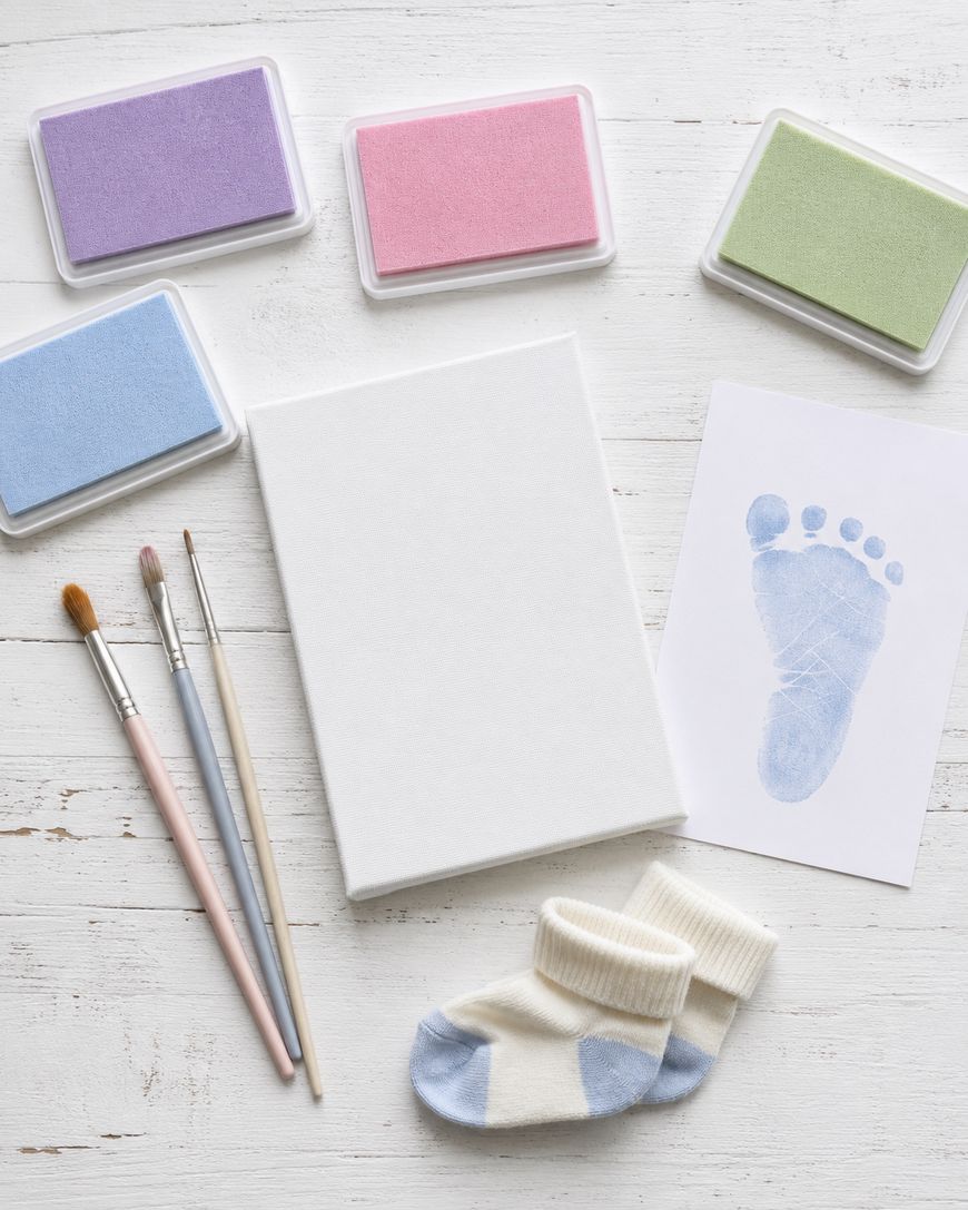 A baby footprint art kit on a white wooden surface, including pastel ink pads, paintbrushes, a blank canvas, a card with a blue footprint, and a pair of tiny socks.