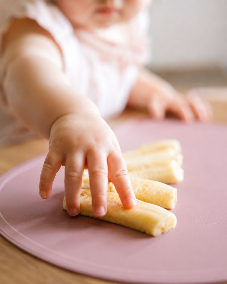 A young child reaching out with their hand to pick up a breadstick from a group of breadsticks on a pink surface.