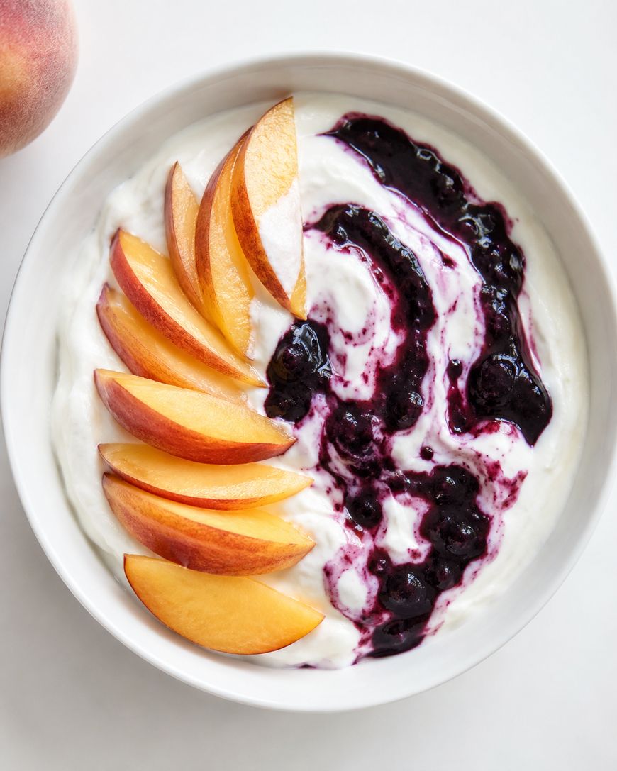 A bowl of yogurt topped with a swirl of blueberry sauce and neatly arranged peach slices on one side, set on a white surface.