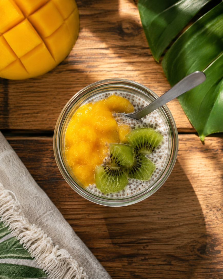 A glass jar of chia pudding topped with mango puree and sliced kiwi sits on a wooden table, next to a napkin, a green tropical leaf, and a partially sliced mango. A spoon is in the jar.
