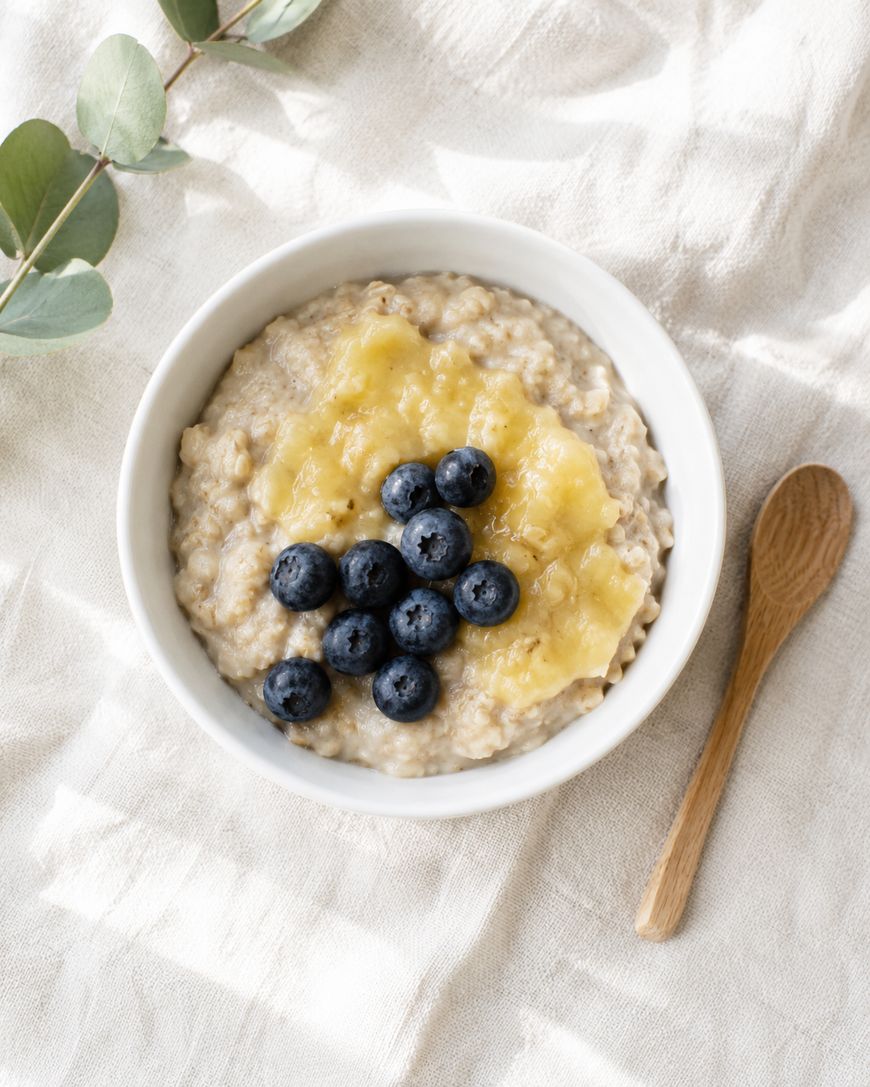 A white bowl of oatmeal topped with mashed banana and fresh blueberries sits on a light fabric surface next to a wooden spoon and a sprig of eucalyptus.