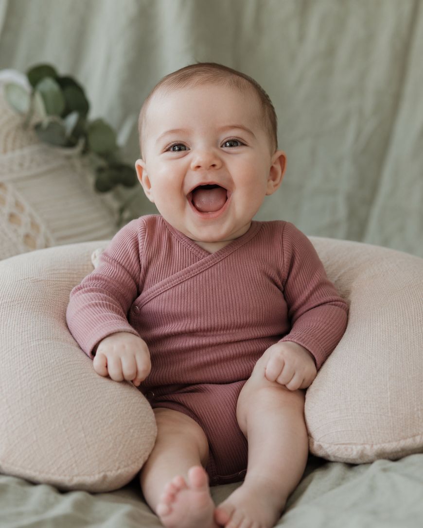 A smiling baby in a mauve onesie sits propped up on a soft, beige cushion, with a green blanket and some leaves in the background.