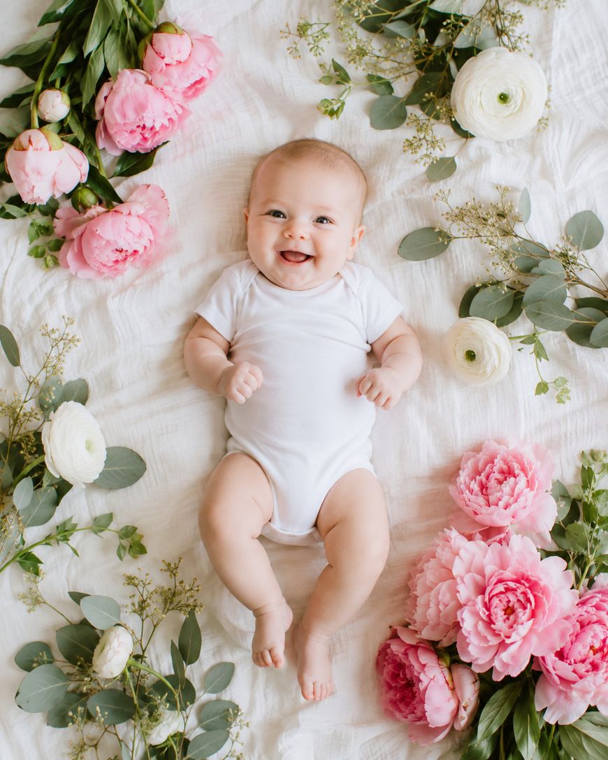 Smiling baby in a white onesie lying on a white blanket, surrounded by pink and white flowers and green leaves arranged in a decorative pattern.