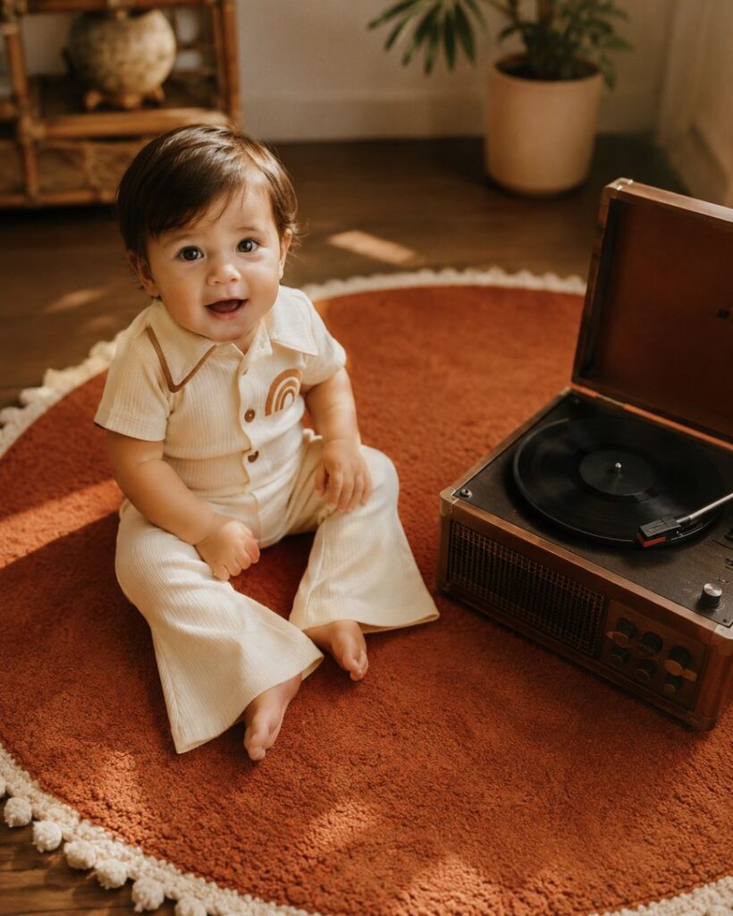 A smiling baby in a cream-colored outfit sits on a round orange rug next to a vintage record player, with warm sunlight streaming into the cozy room decorated with plants.