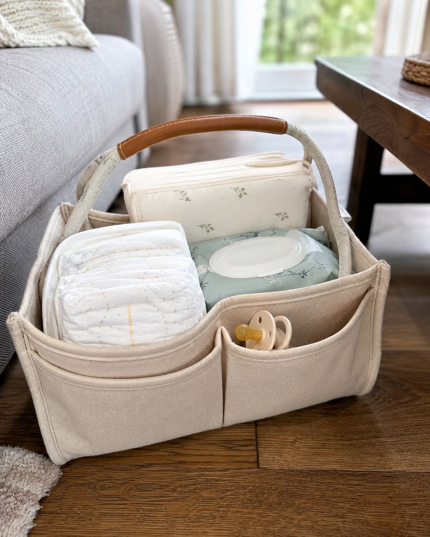 A beige fabric caddy with a leather handle sits on a wooden floor. It holds diapers, baby wipes, a pacifier, and a floral-patterned pouch, placed next to a sofa and a wooden table in a bright room.