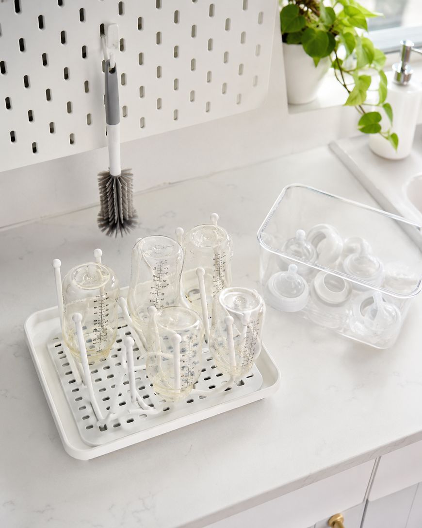 Four clear baby bottles drying upside down on a white rack, with bottle parts in a plastic container next to them on a white kitchen counter near a sink and potted plant.