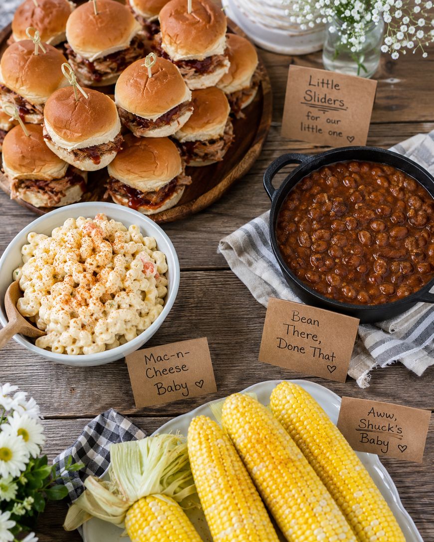 A rustic table displays sliders, macaroni and cheese, baked beans, and corn on the cob, each with handwritten cards featuring playful baby-themed puns. White flowers decorate the corners of the table.
