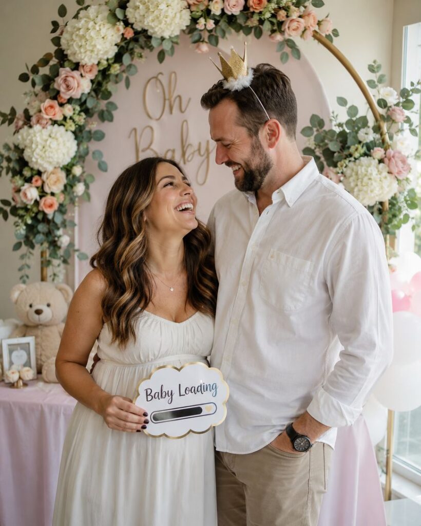A smiling pregnant woman in a white dress holds a “Baby Leading” sign next to a man wearing a gold crown. They stand in front of a pink floral baby shower backdrop with “Oh Baby” written on it.