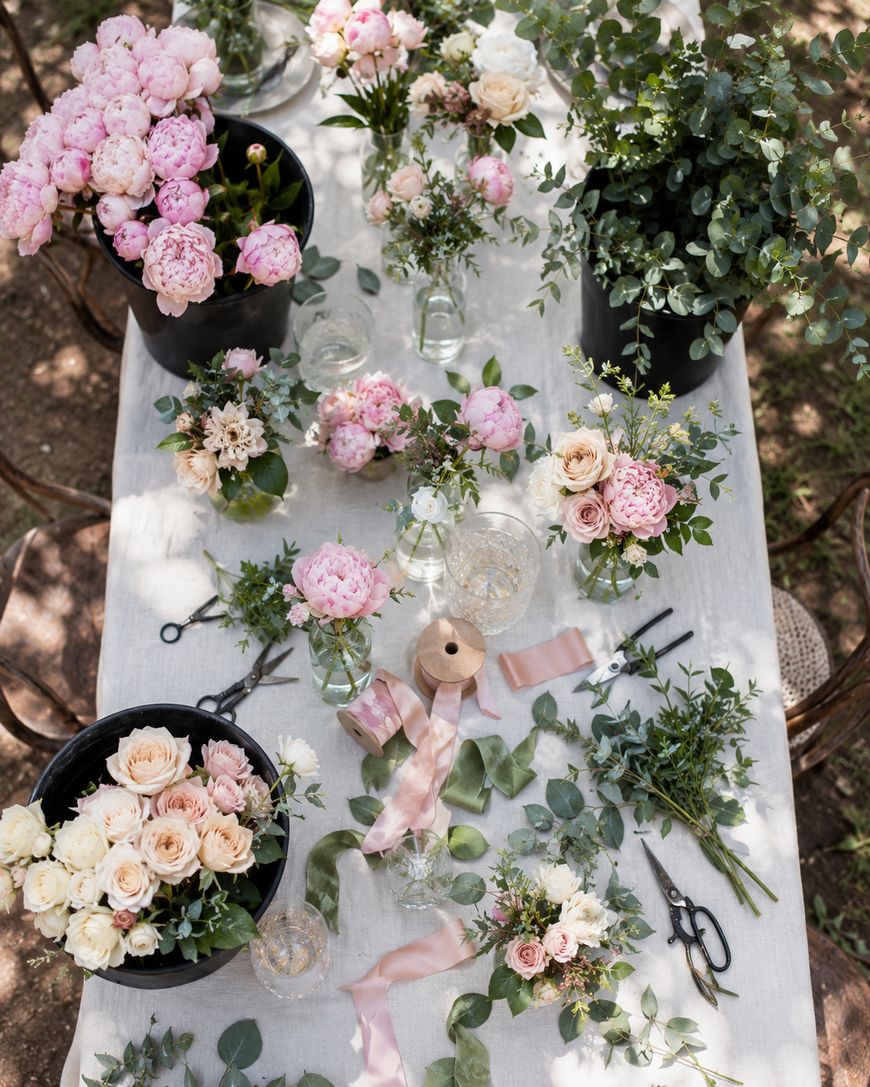A top view of a table decorated with pink peonies, white roses, greenery, floral vases, ribbon, scissors, and loose leaves, creating a lush, romantic floral arrangement scene outdoors.
