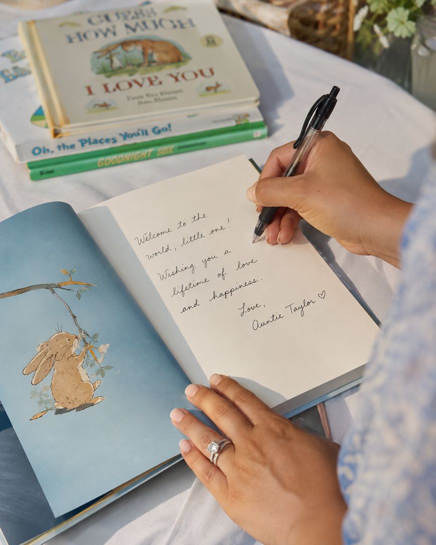 A person writes a heartfelt note in a children’s book with an illustration of a bunny. Stacked books, including Goodnight Moon and Oh, the Places You’ll Go!, sit on a white table in the background.
