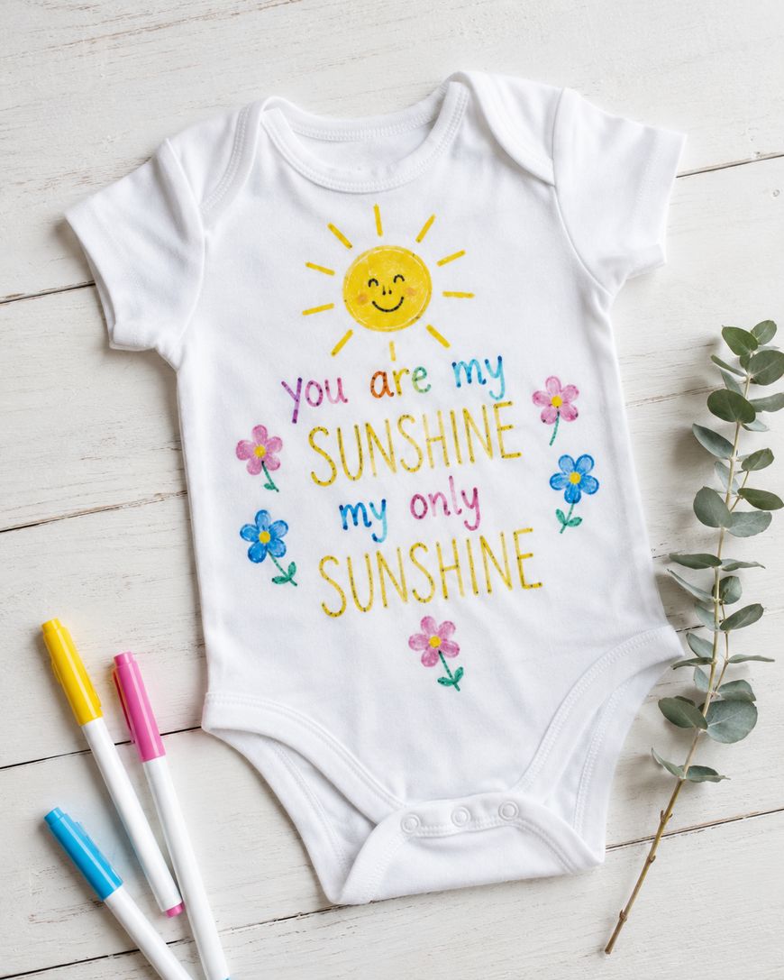 A white baby onesie with colorful embroidery that reads You are my sunshine, my only sunshine with a smiling sun and flowers. Three colorful markers and a sprig of greenery lie nearby on a white wooden surface.
