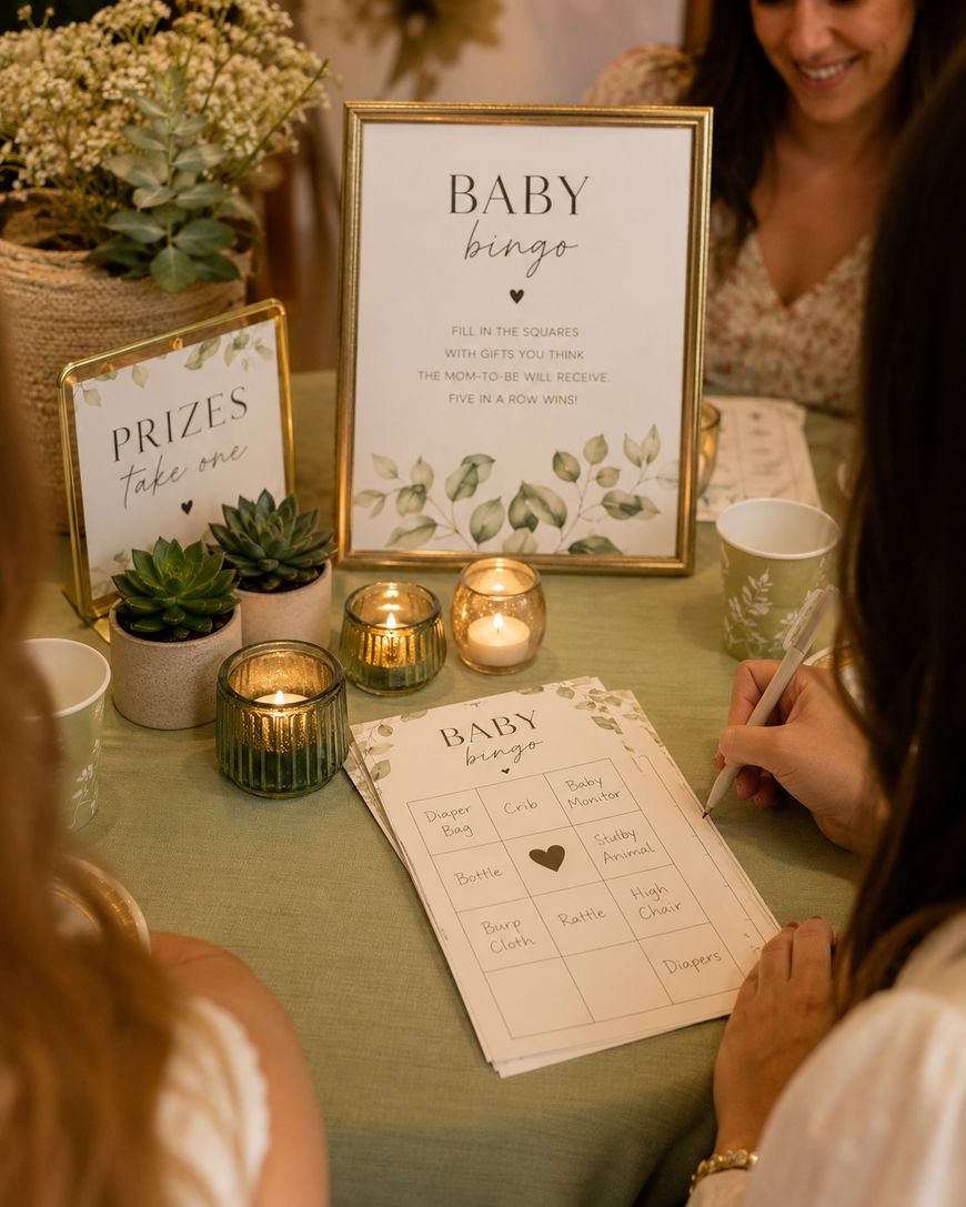 Two women sit at a green table playing baby shower bingo, surrounded by candles, potted plants, and a Prizes take one sign. One woman writes on a bingo card; a Baby Bingo instruction sign is displayed upright.