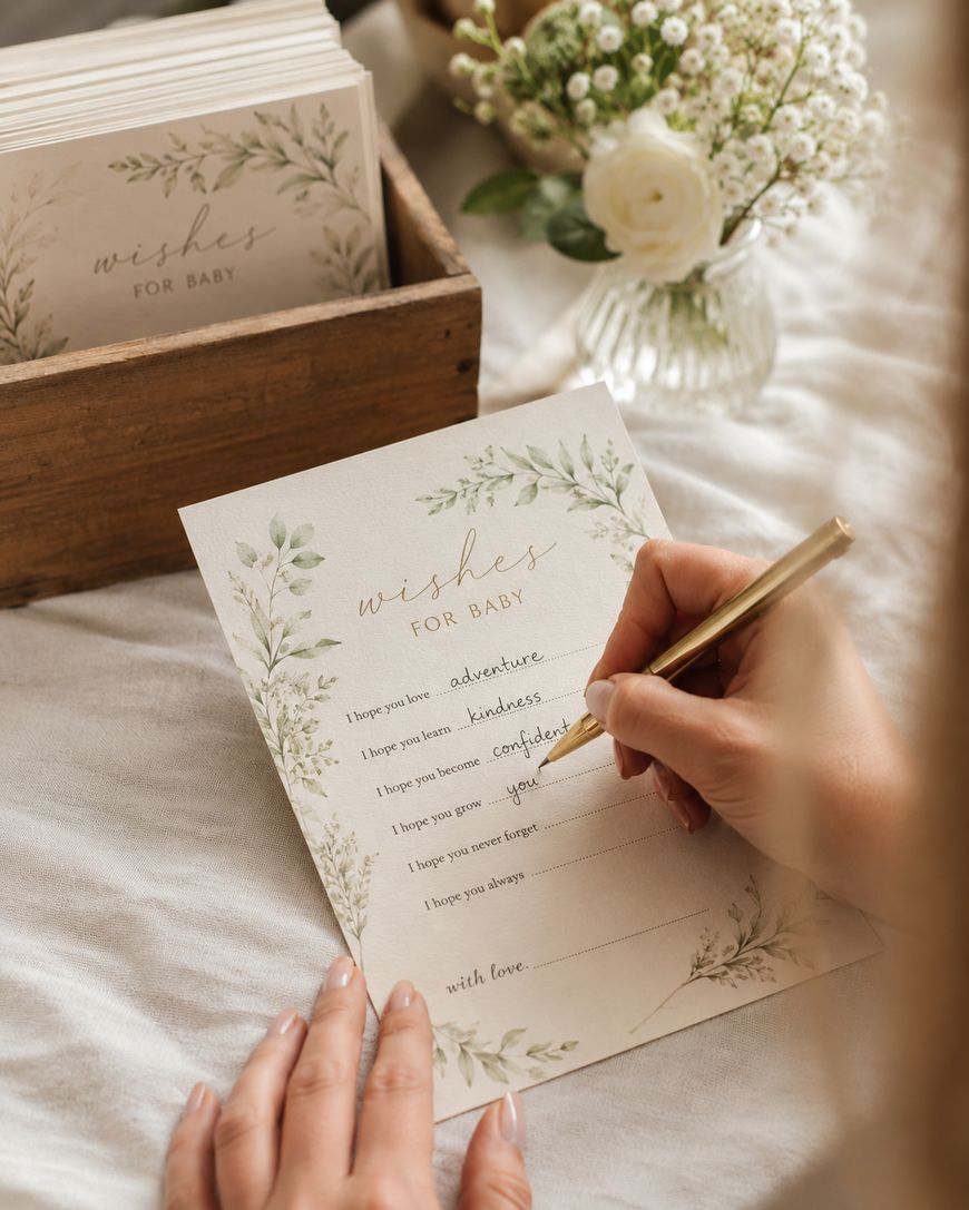 A person writes wishes for a baby on a decorative card with floral designs. Other similar cards are in a wooden box nearby, and a small vase with white flowers is in the background.