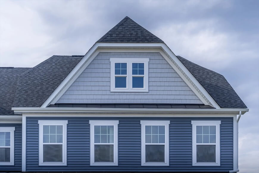 The upper portion of a blue two-story house with gray shingles, white trim, and multiple windows, photographed under a cloudy sky.