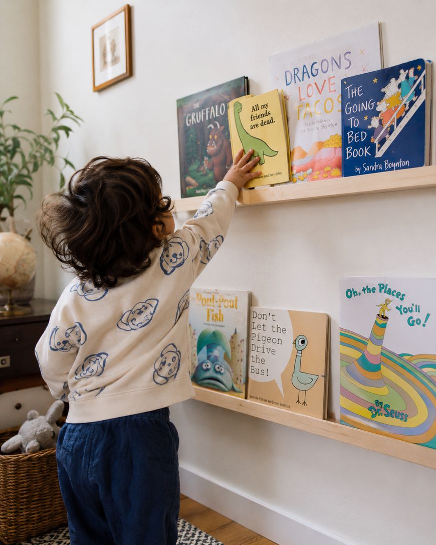 A young child in a sweater with bear faces reaches for a book on a low wooden bookshelf filled with colorful children’s books in a cozy, well-lit room.