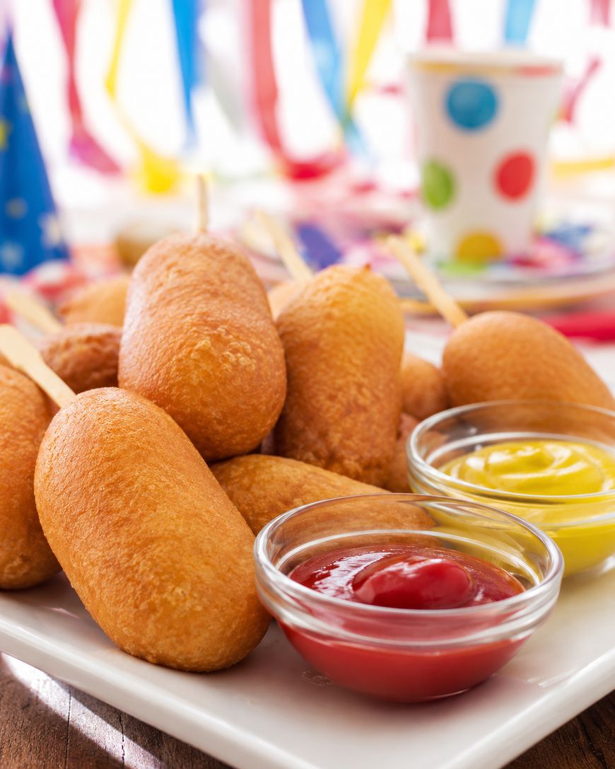 A plate of corn dogs on sticks is served with small bowls of ketchup and mustard. Colorful party decorations and a paper cup are blurred in the background.
