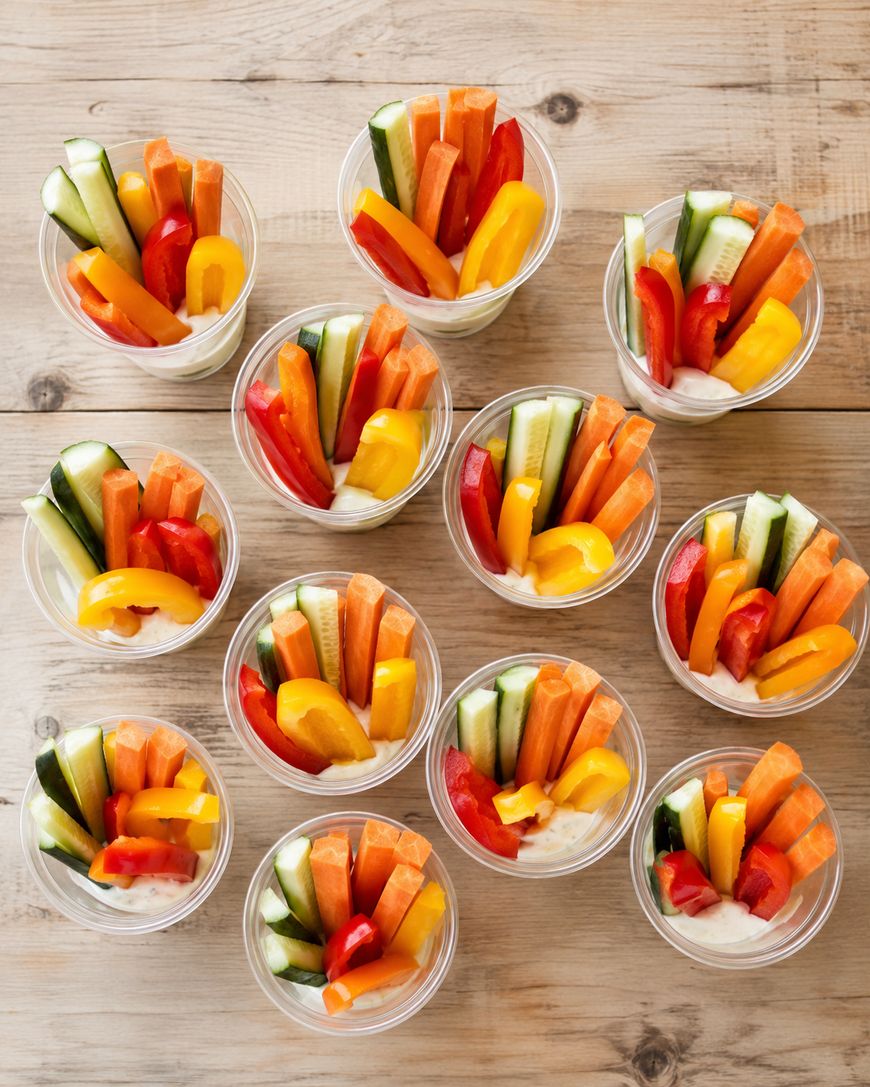Twelve clear cups filled with sliced cucumbers, carrots, red and yellow bell peppers, and a white dipping sauce, arranged on a wooden surface.