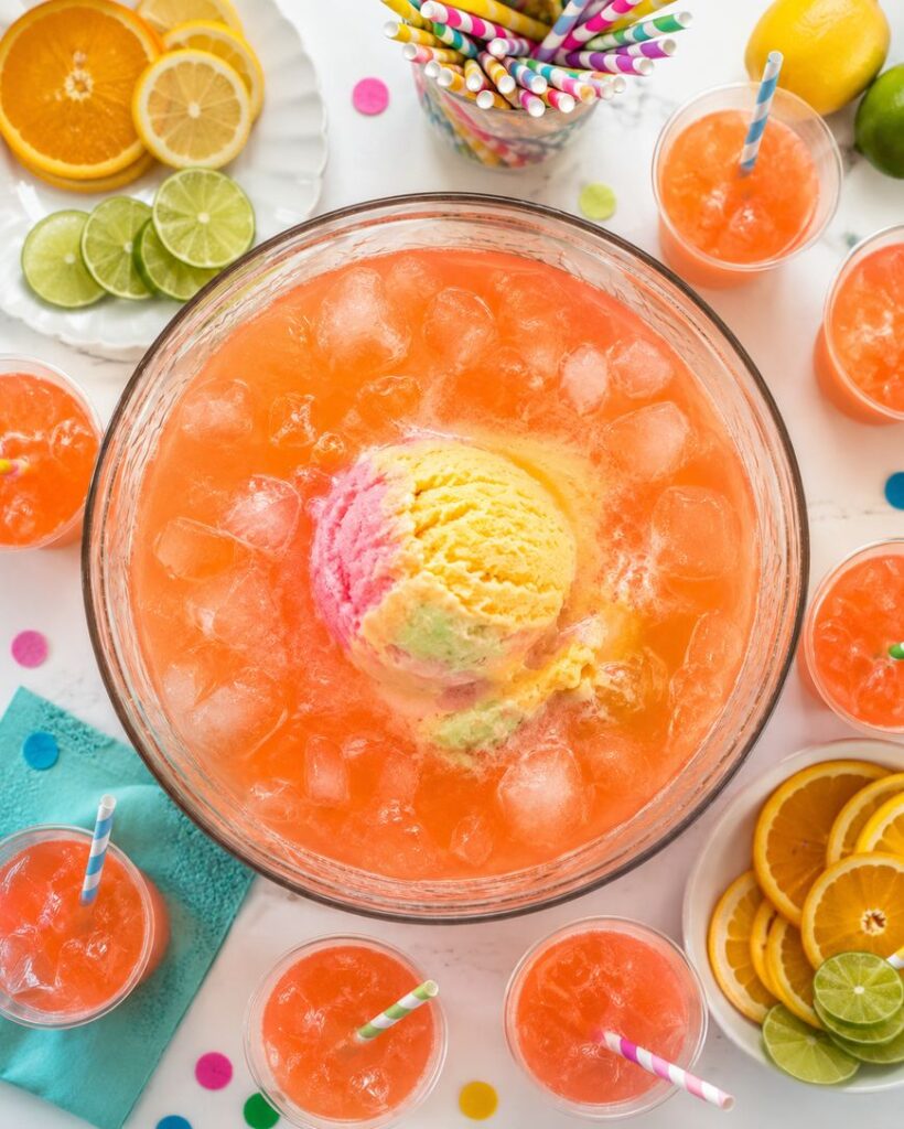 A large glass bowl of orange punch with ice and a scoop of rainbow sherbet in the center, surrounded by glasses of punch, colorful straws, and sliced citrus fruits on a bright, festive table.