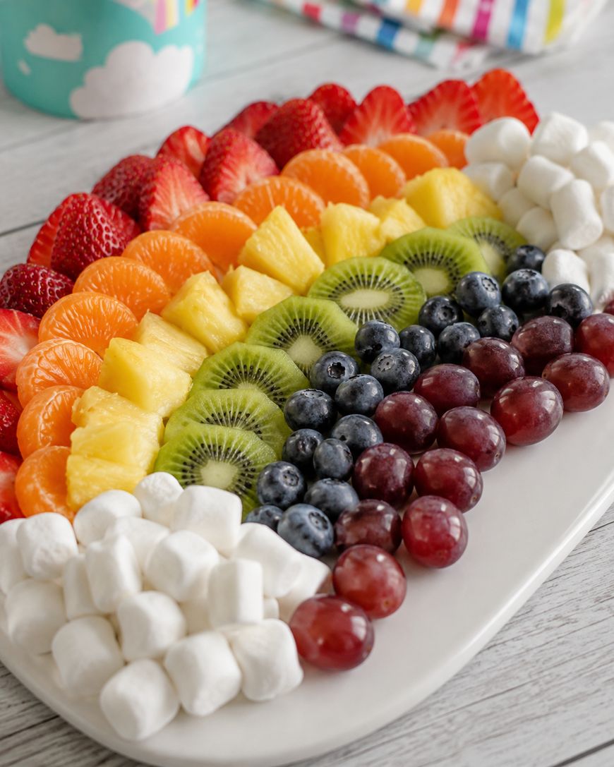 A white platter arranged with rows of fruit and marshmallows in a rainbow pattern: strawberries, mandarin oranges, pineapple, kiwi, blueberries, red grapes, and mini marshmallows. A colorful napkin is in the background.