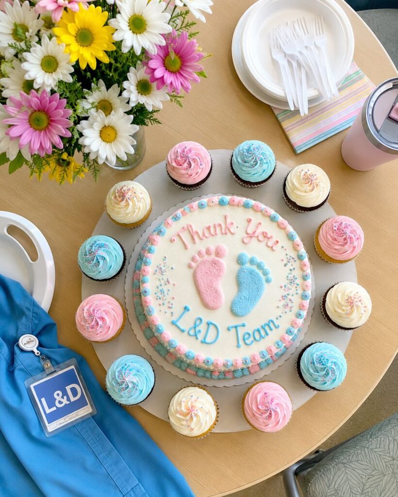 A round cake with pink and blue baby footprints and Thank You L&D Team written on top is surrounded by pink and blue frosted cupcakes. Nearby are flowers, utensils, a pink cup, and a blue L&D badge on scrubs.