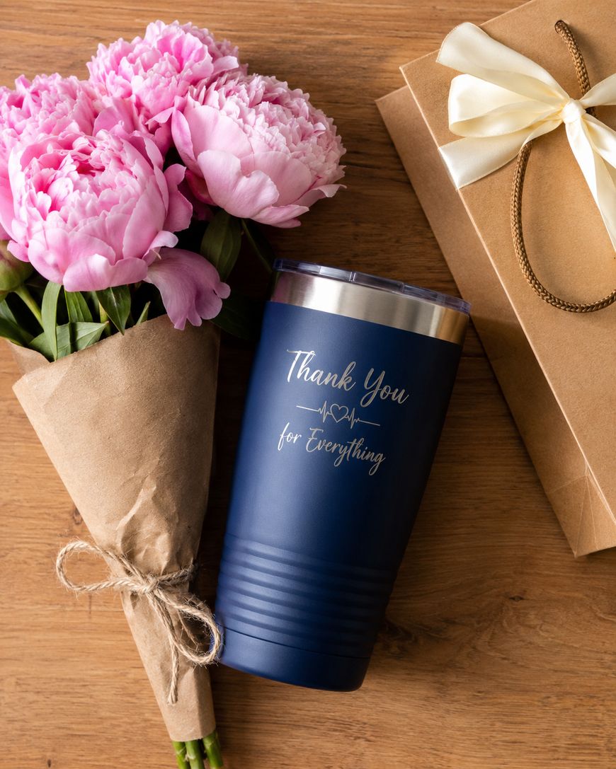 A bouquet of pink flowers, a navy blue tumbler with Thank You for Everything written on it, and a gift box with a cream ribbon, all arranged on a wooden surface.