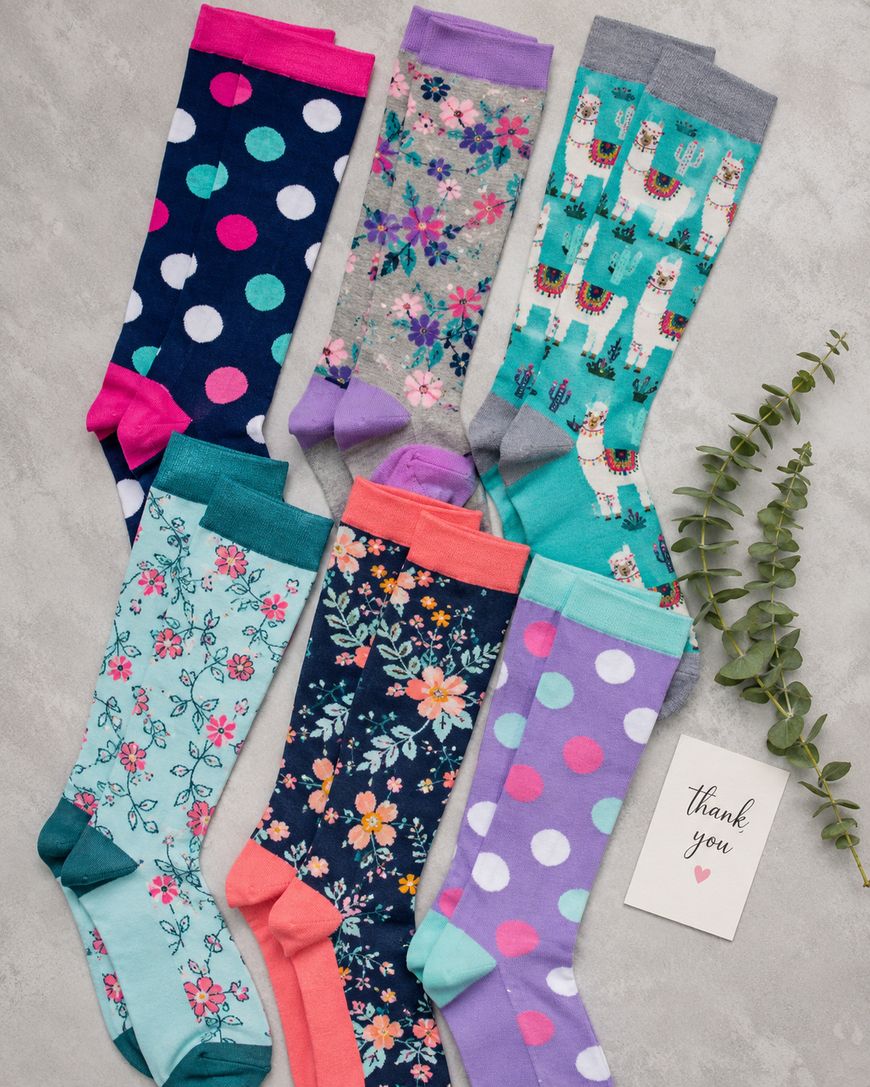 Seven pairs of colorful socks with various patterns, including polka dots, florals, and llamas, are arranged on a gray surface next to a sprig of greenery and a small card that says thank you.
