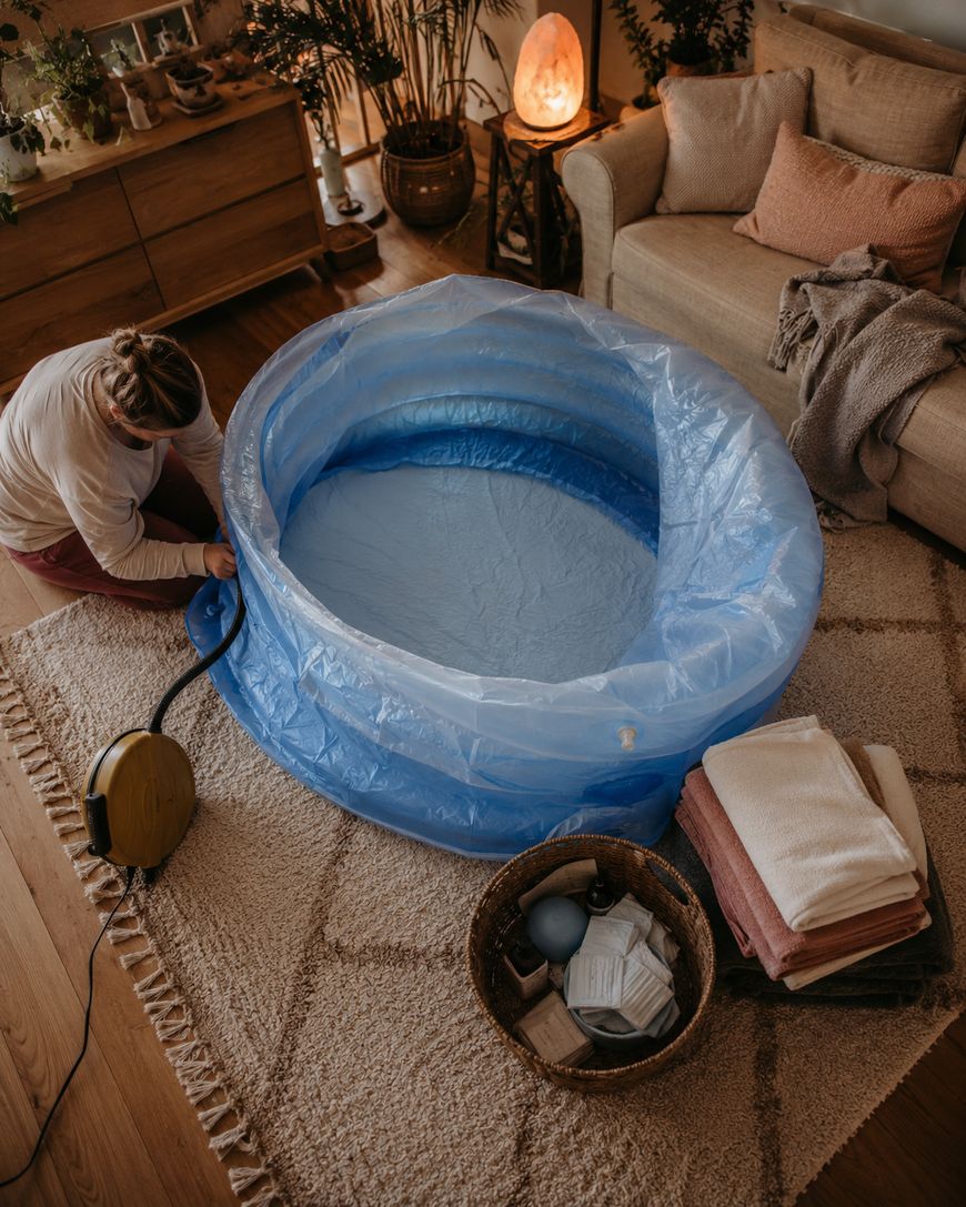 A person inflates a blue birthing pool with an air pump in a cozy living room. Nearby are a basket with supplies and folded towels on a beige rug. The room has warm lighting and comfortable furnishings.