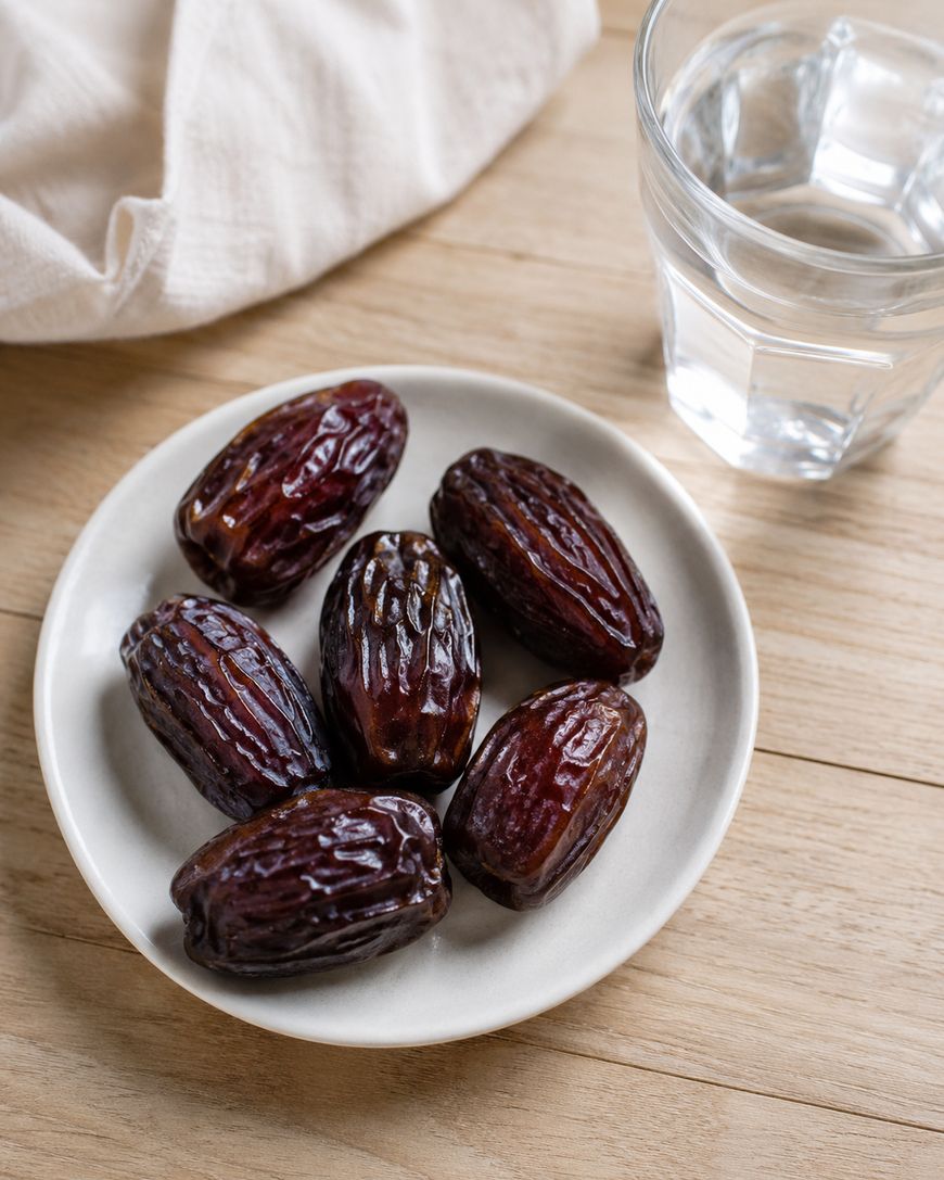 A small white plate with six glossy dates sits on a wooden table next to a glass of water and a light-colored cloth.