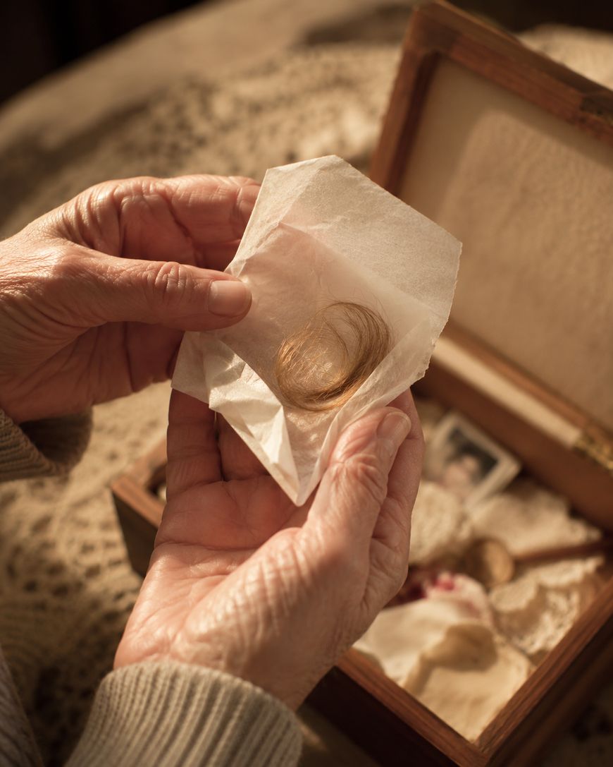 An elderly person holds a lock of light brown hair wrapped in tissue paper, with a wooden memory box containing old photographs and keepsakes in the background.