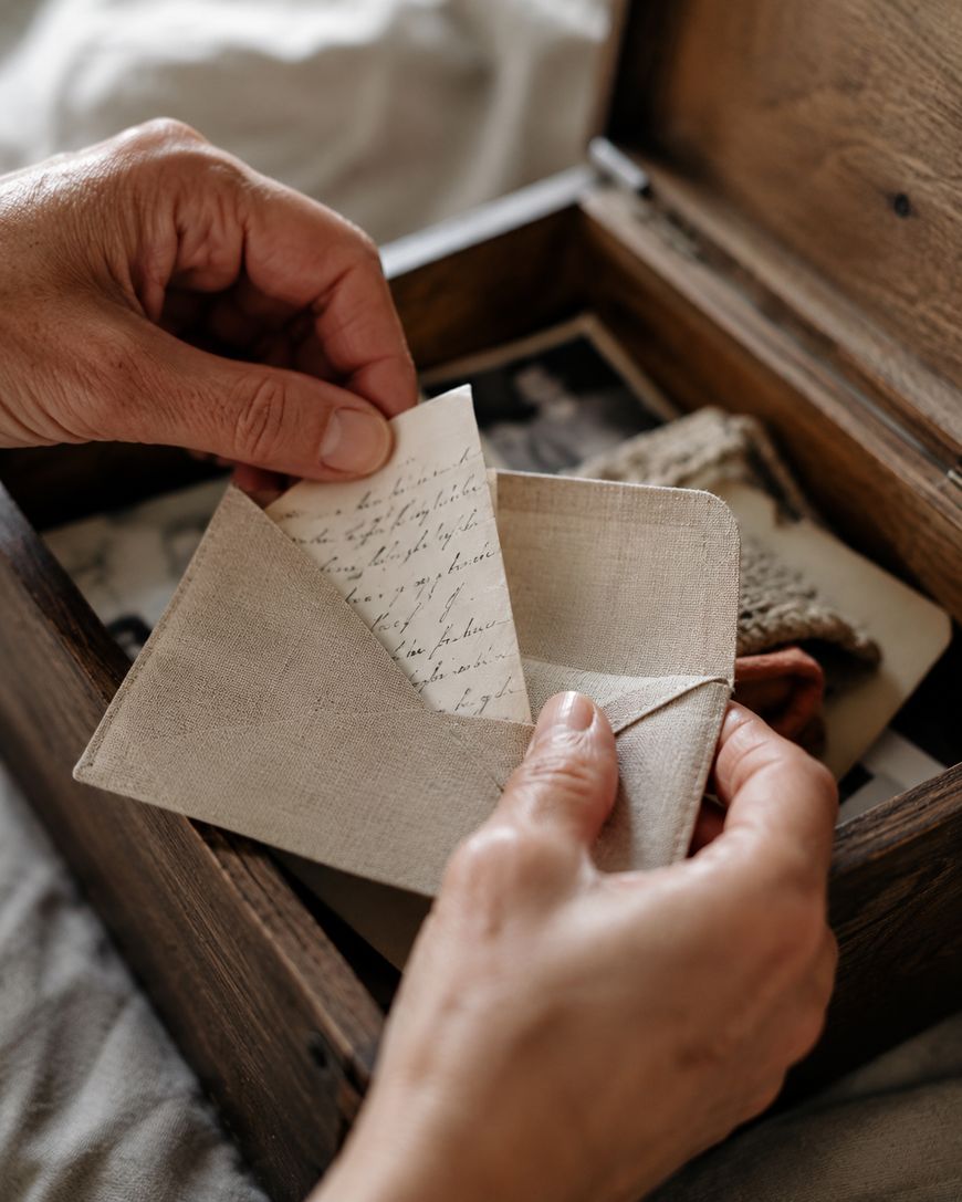 A person opens a fabric envelope containing a handwritten letter, with a wooden box filled with keepsakes and photographs in the background.