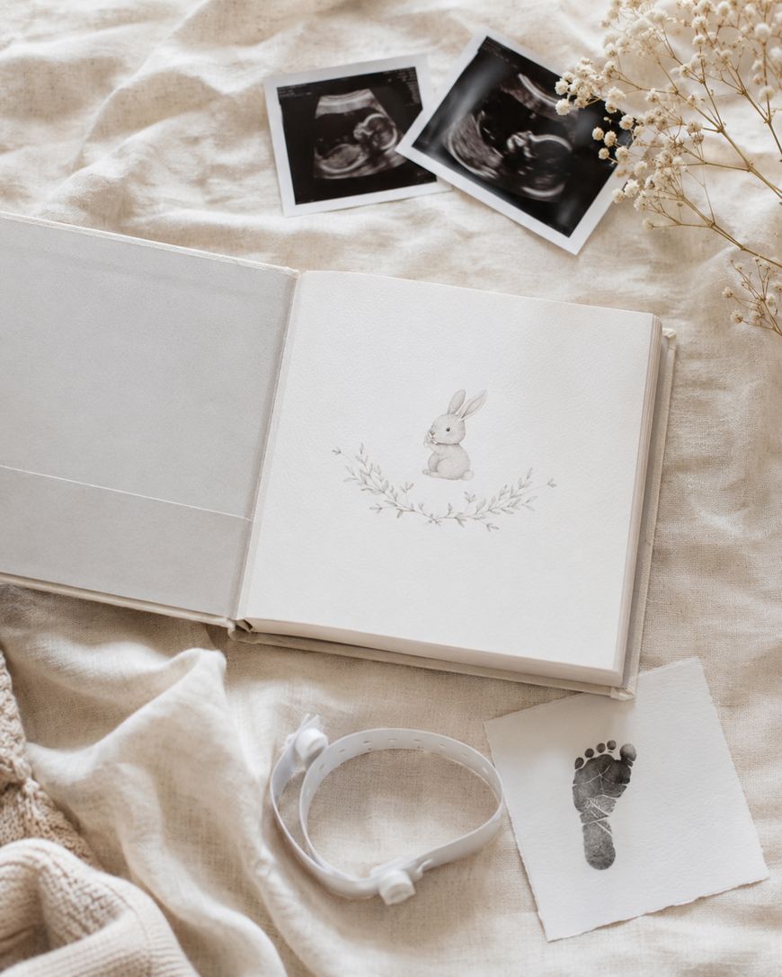 An open baby memory book with a bunny illustration, two ultrasound photos, a dried flower, a hospital ID bracelet, and a paper with a baby’s footprint, all arranged on a soft beige fabric.