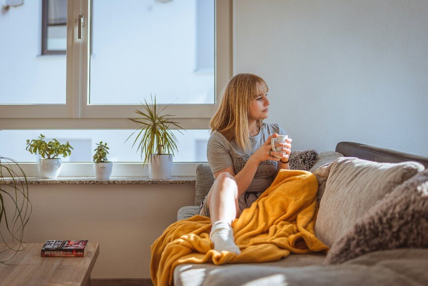 A woman sits on a couch with a yellow blanket, holding a mug and looking out a large window. There are potted plants on the windowsill and sunlight fills the cozy room.