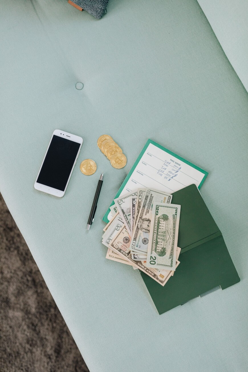A smartphone, a pen, gold coins, a bill payment receipt, and a green folder with assorted US dollar bills are arranged on a light blue surface.