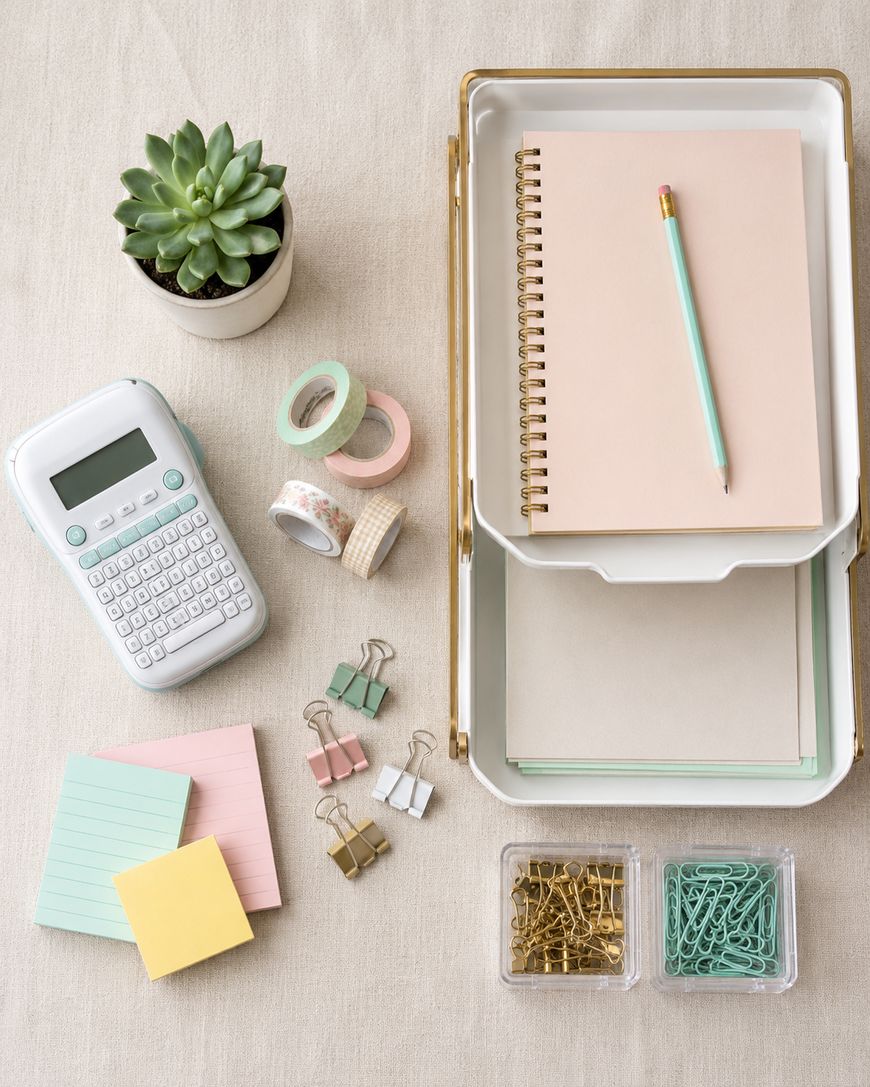 A neatly organized desktop with a potted succulent, a label maker, pastel notepads, binder clips, gold paper clips, green paper clips, decorative tape rolls, and stacked notebooks with a pencil on a beige surface.