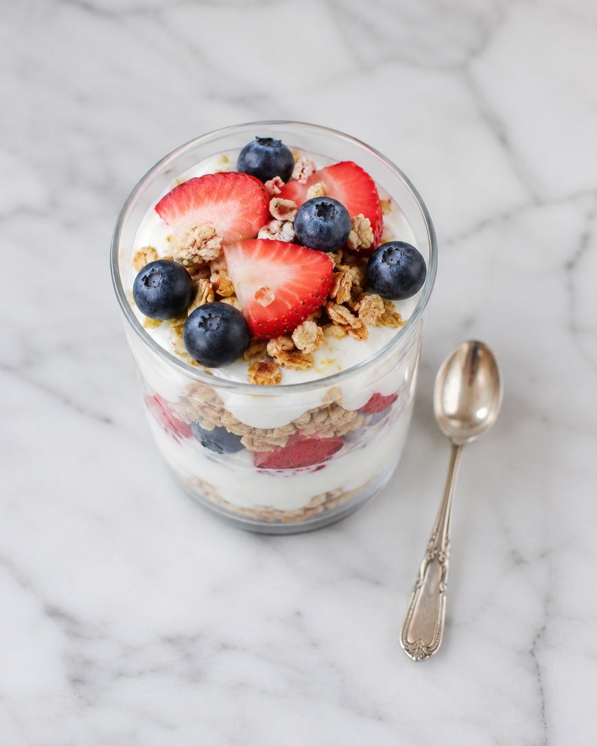 A glass filled with layers of yogurt, granola, strawberries, and blueberries sits on a marble surface next to a silver spoon.