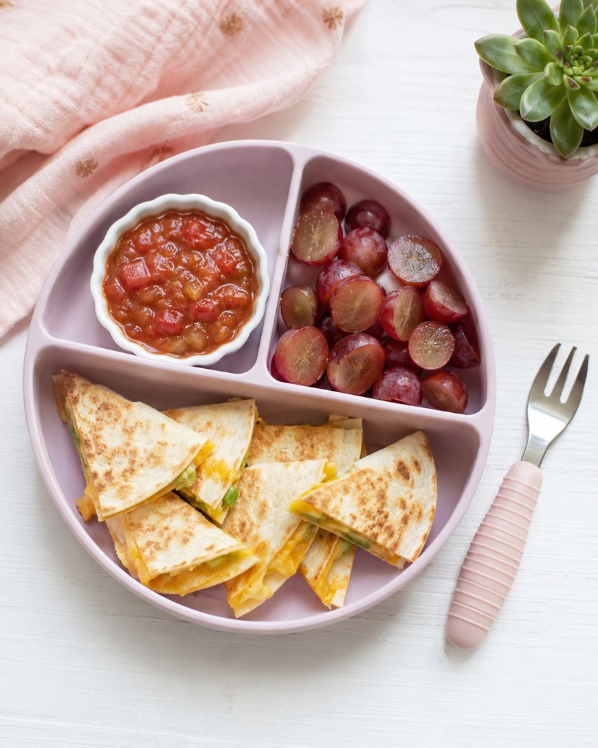 A pink plate with cheese quesadilla slices, halved red grapes, and a small dish of salsa. A pink fork and a green succulent plant are also visible on a white table.