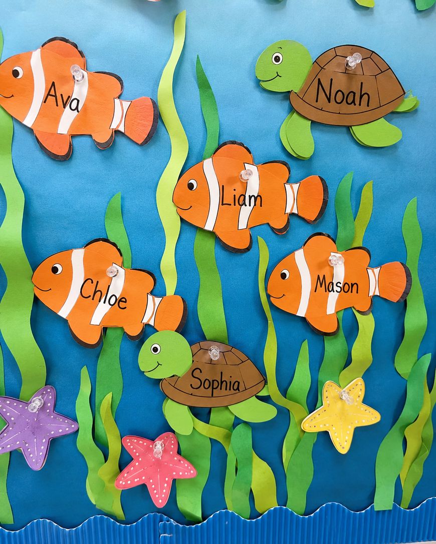 Colorful classroom bulletin board with paper cutouts of orange clownfish and green turtles, each labeled with names: Ava, Liam, Chloe, Mason, Noah, and Sophia. Green seaweed and starfish decorations complete the underwater theme.