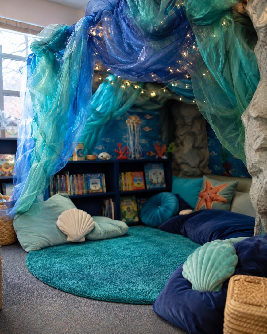 A cozy reading nook with an under-the-sea theme, featuring a blue-green canopy with string lights, shell-shaped pillows, plush seating, an aqua rug, bookshelves, and ocean-themed decor.